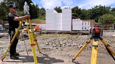 En man står på en byggplats vid ett stativ med mätverktyg. Han är klädd i svart keps och svarta kläder.