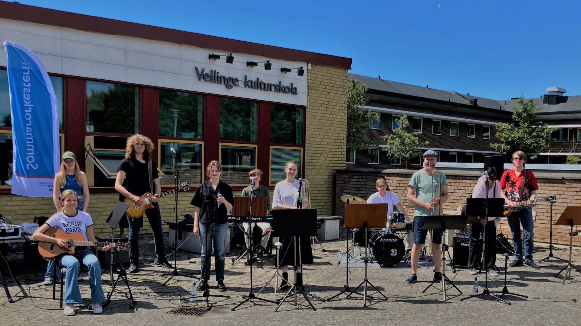 Ungdomar och vuxna sitter eller står med varsitt musikinstrument framför en gul tegelbyggnad med ordet Kulturskolan på väggen. Solen skiner och alla ler mot kameran.