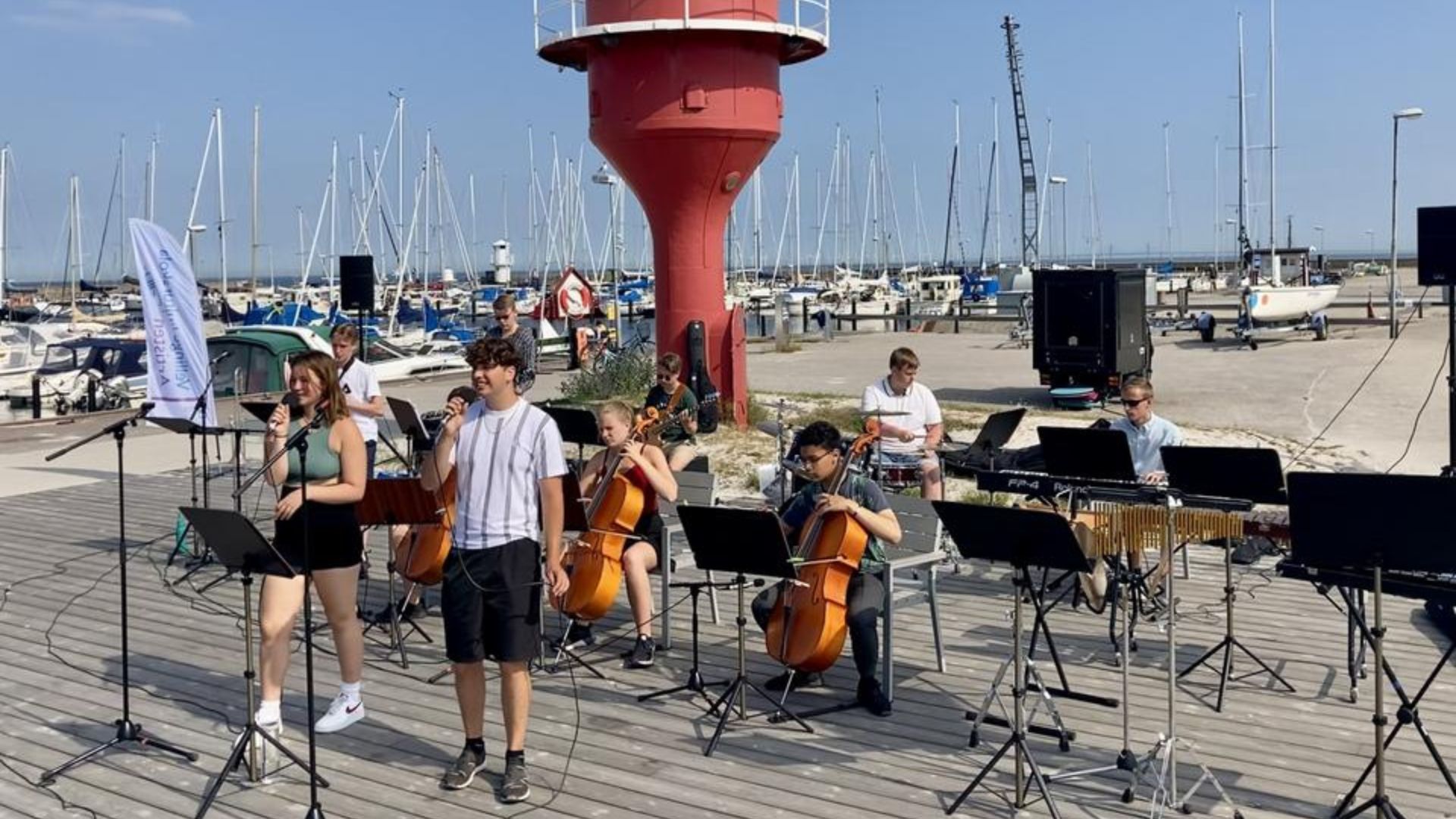 Ungdomar spelar musik och sjunger i Skanörs hamn en solig dag. De står på ett trädäck och i bakgrunden syns småbåtar i hamnen.