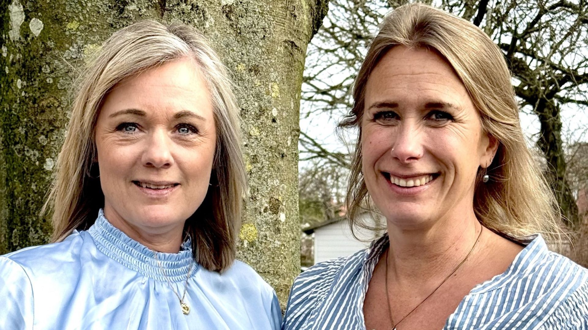 Två kvinnor står bredvid varandra framför ett träd. Den ena har en ljusblå satinblus och blont hår. Den andra har en blå- och vitrandig blus och mellanblont hår. 