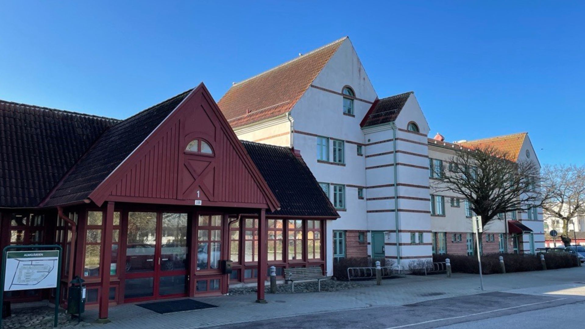 Entré i rött trä. Intill ligger ett trevåningshus i vit putsad fasad med orange tegeltak.
